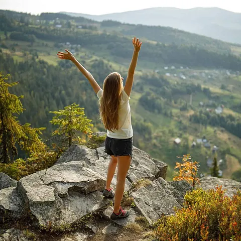 Kvinne står på fjellkant med armene hevet og ser ut over et grønt dalføre med skog og spredte hus.